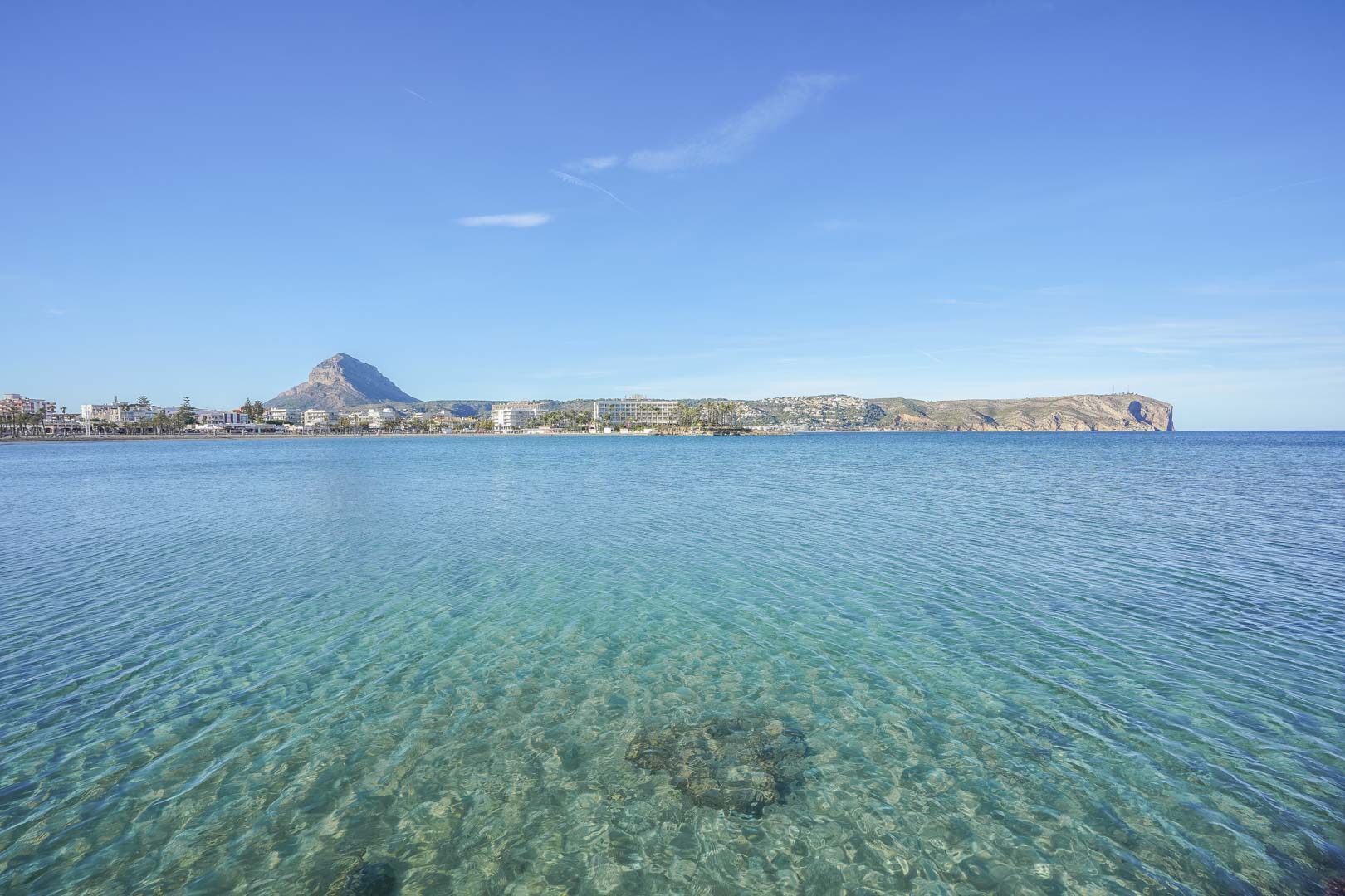 Vue panoramique sur la baie de Jávea aux eaux turquoises cristallines, avec le mont Montgó et les falaises du Cabo de San Antonio à l'horizon.