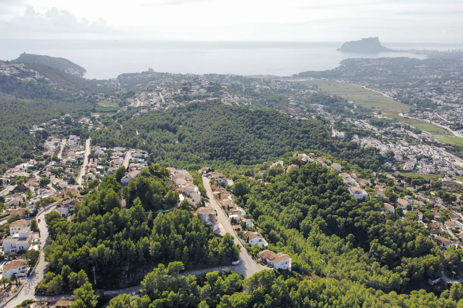 Vue aérienne panoramique du quartier de Paichi, illustrant la forêt de pins, les villas avec piscines et le Peñón d'Ifach se découpant sur l'horizon méditerranéen.