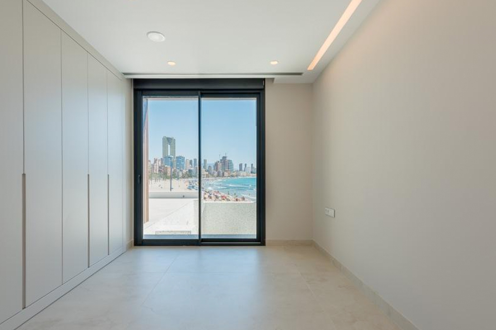 Chambre avec placards intégrés, éclairage LED encastré et baie vitrée donnant sur la plage de Poniente et les gratte-ciel de Benidorm.