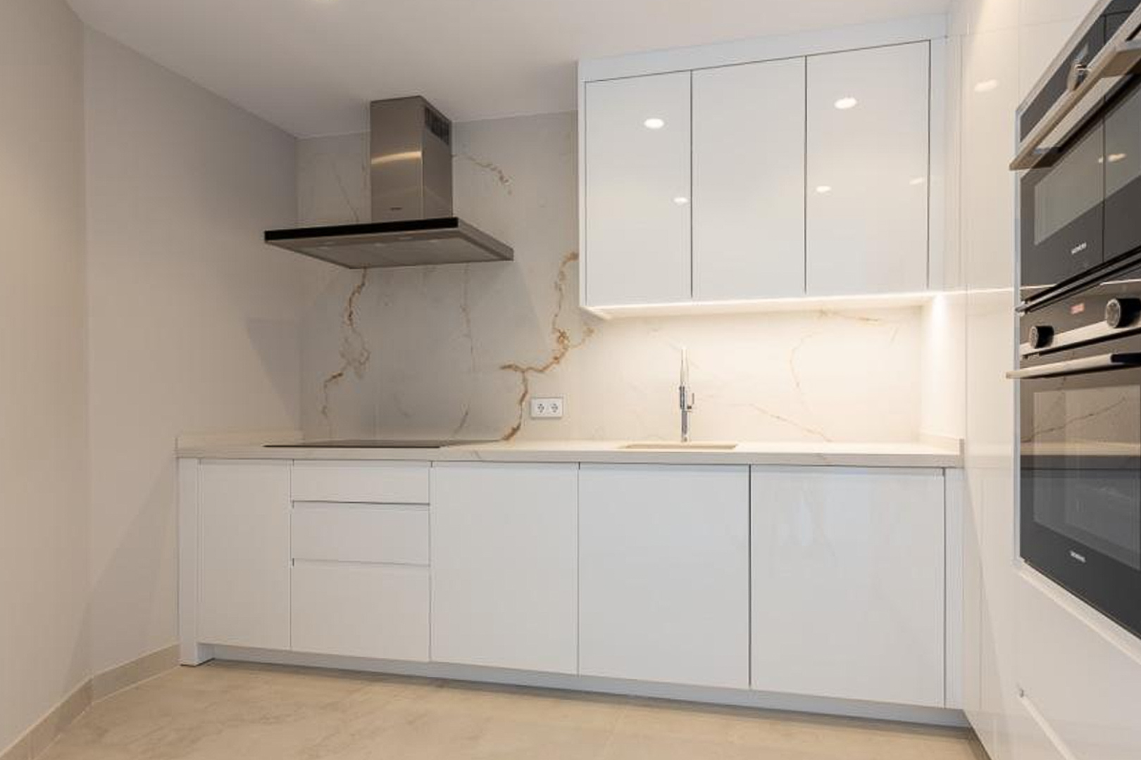 Cuisine minimaliste avec placards blancs sans poignées, crédence en pierre marbrée et électroménagers encastrés haut de gamme sous un éclairage LED intégré.