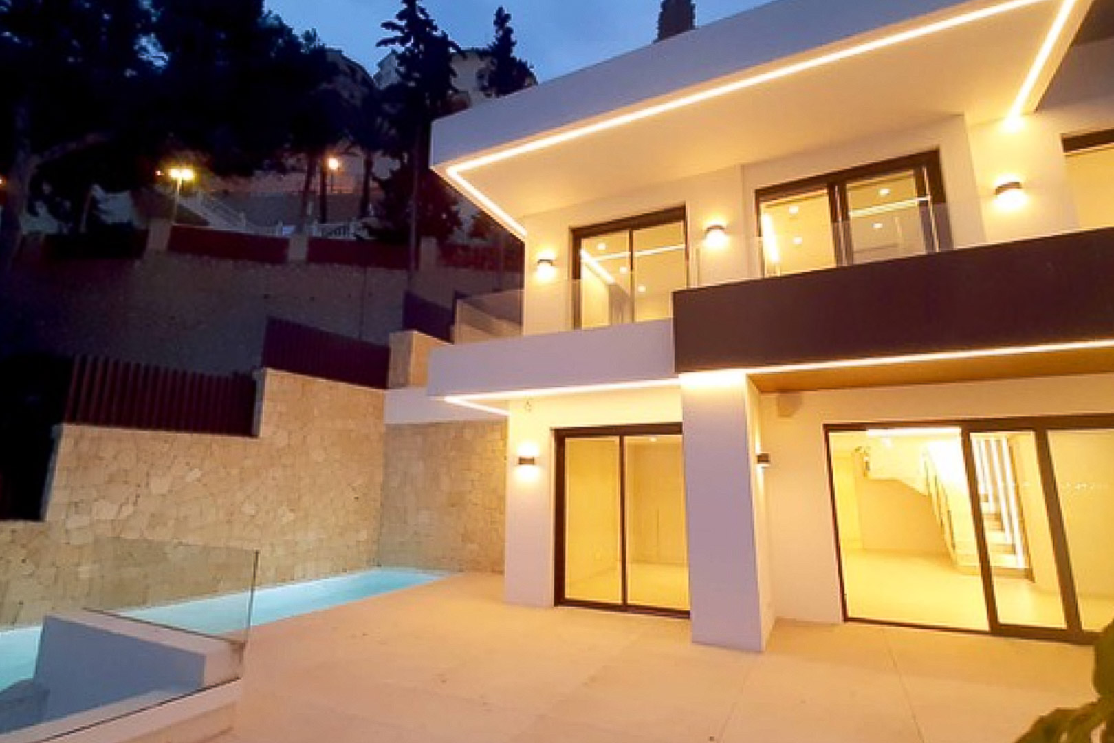 Façade moderne à deux niveaux avec éclairage LED intégré, grandes baies vitrées et terrasse avec piscine privée bordée d'un mur en pierre naturelle.