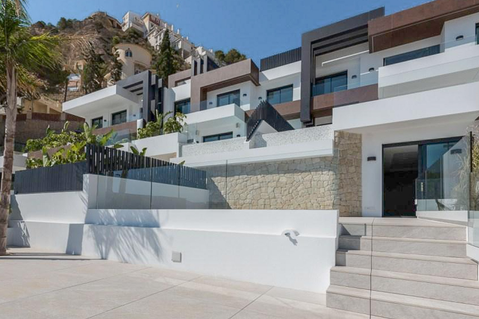 Façade moderne à niveaux décalés avec murs en pierre naturelle, grandes terrasses aux garde-corps en verre et escalier d'accès en grès cérame.
