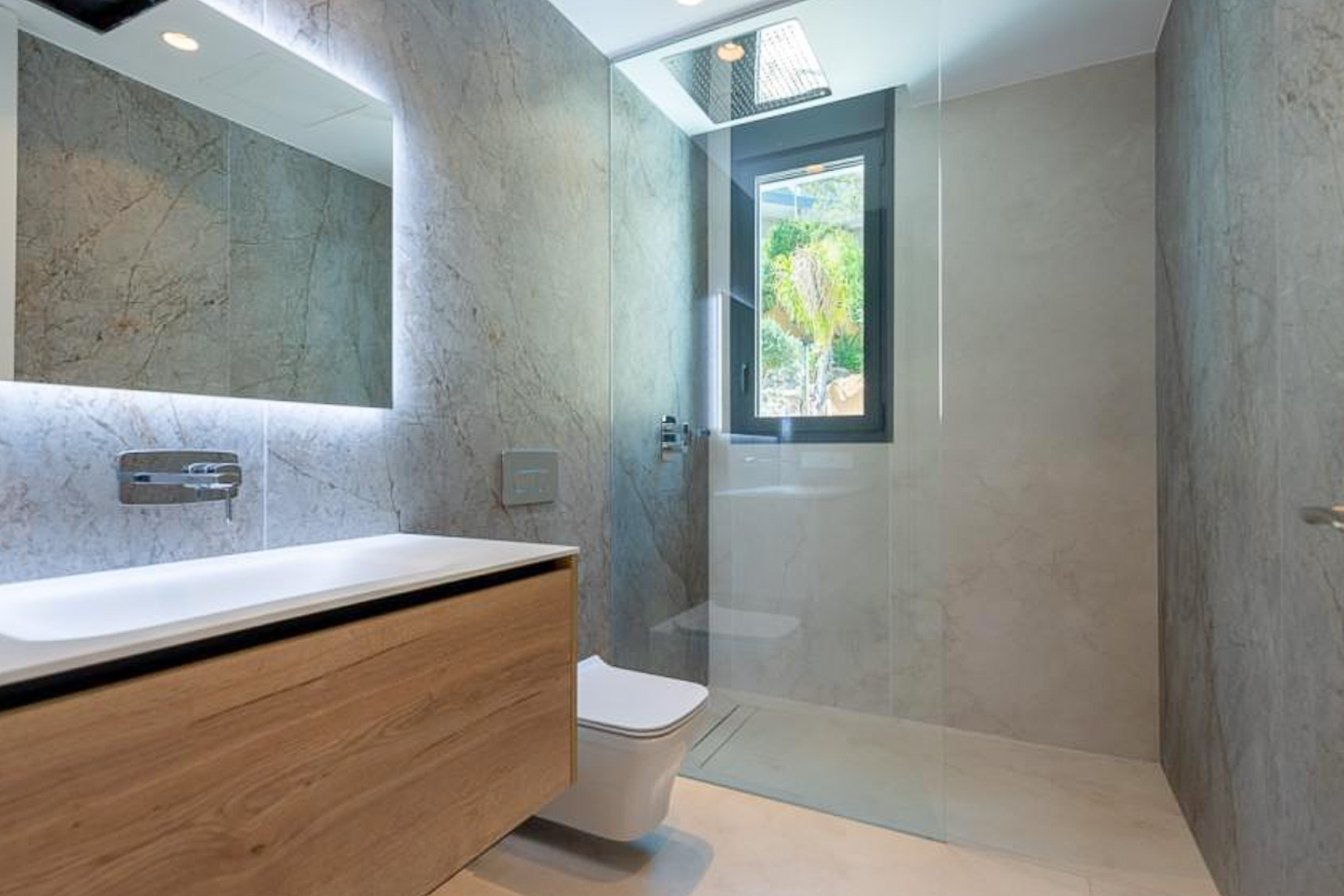 Salle de bain design avec carrelage effet pierre, meuble vasque suspendu en bois, miroir rétroéclairé et douche à l'italienne avec ciel de pluie intégré.