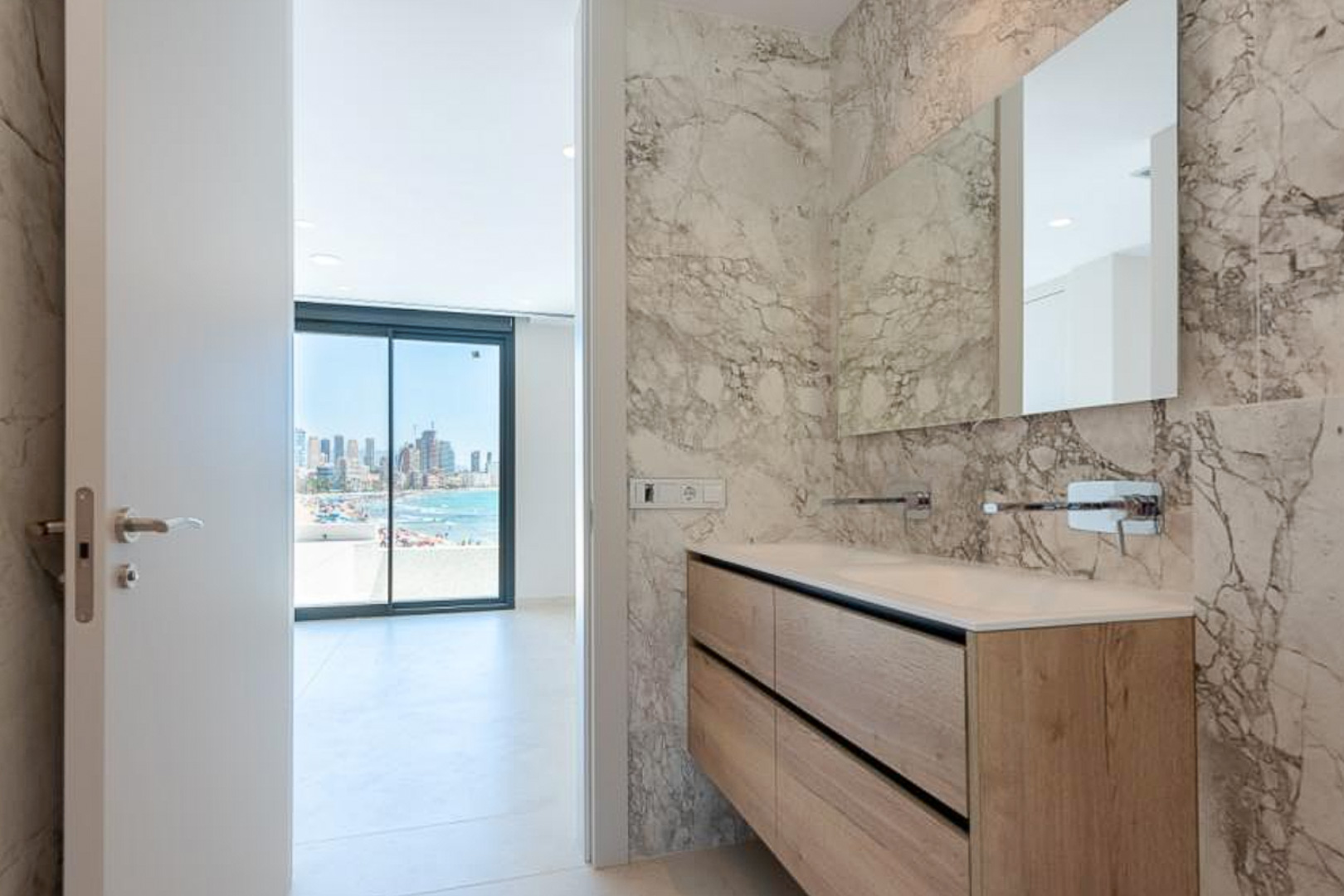 Salle de bain élégante avec murs en marbre, meuble vasque double en bois et robinetterie murale, offrant une perspective sur la mer.