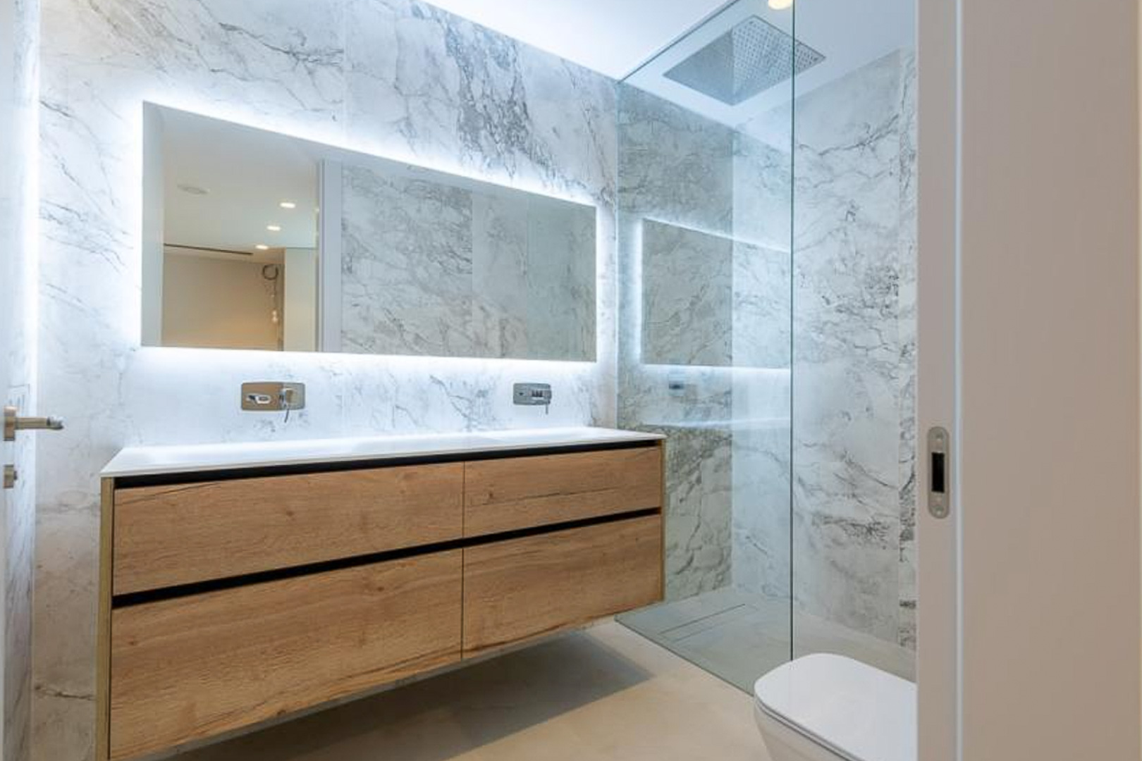 Salle de bain moderne avec carrelage effet marbre, meuble double vasque suspendu en bois et miroir rétroéclairé. Douche à l'italienne avec paroi vitrée.