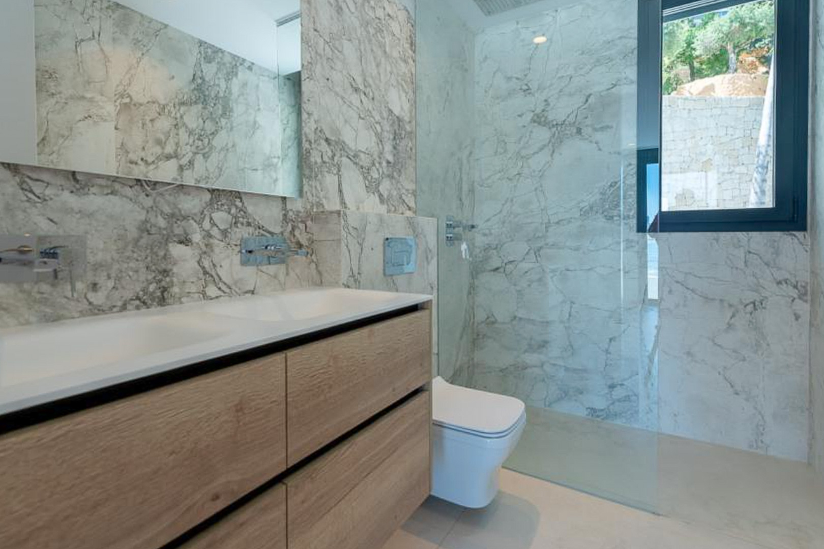 Salle de bain moderne avec revêtement en marbre, meuble double vasque en bois clair et douche à l'italienne avec paroi en verre fixe.