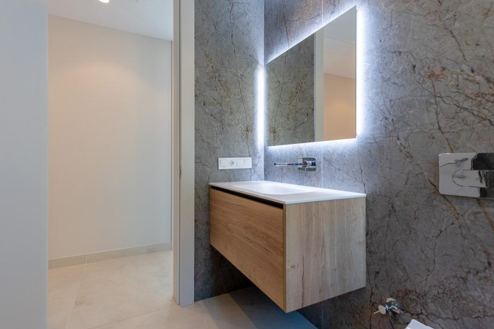 Salle d'eau moderne avec meuble suspendu en bois, vasque intégrée et miroir rétroéclairé sur un mur en grès cérame effet marbre gris.