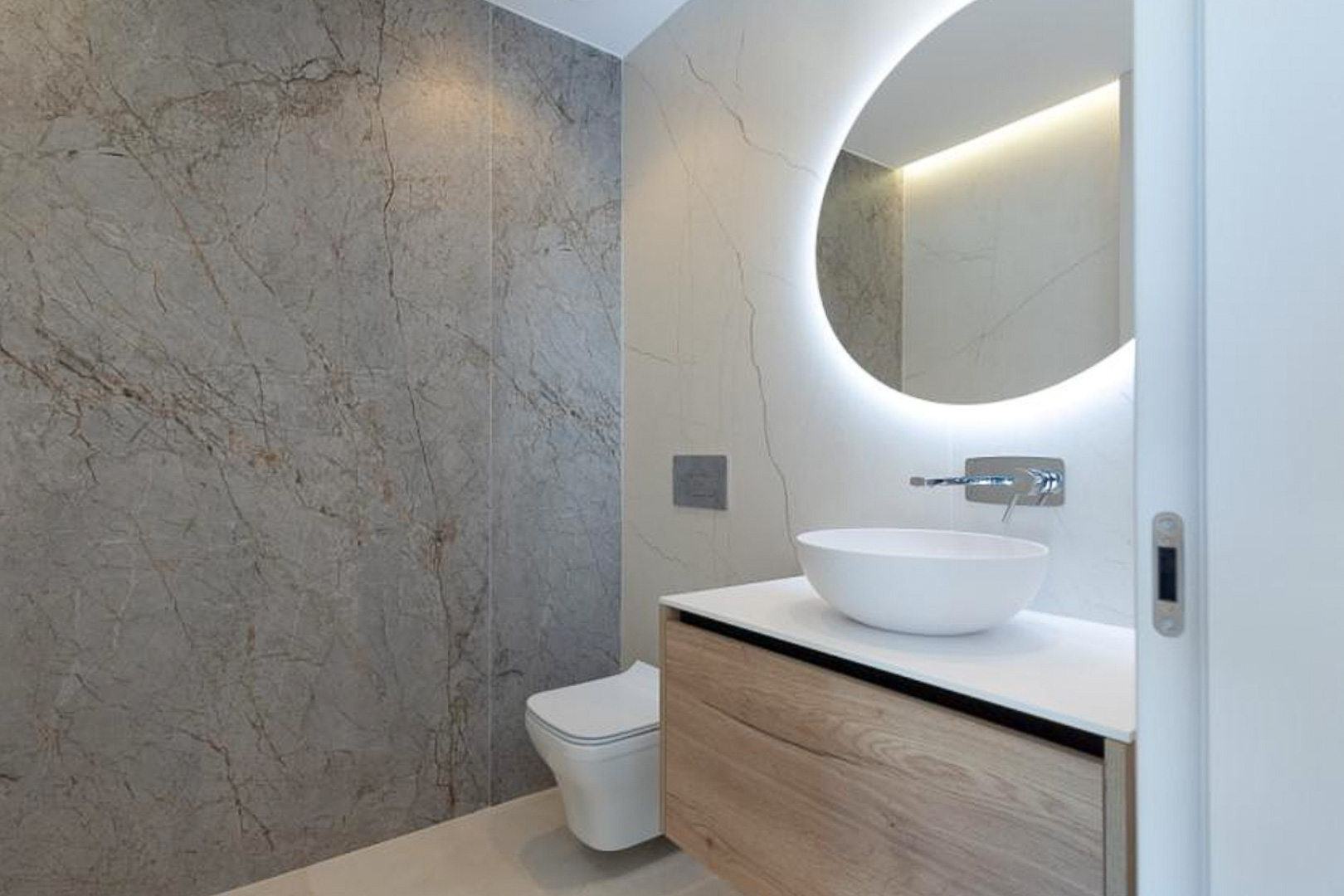 Salle d'eau moderne avec meuble vasque suspendu en bois, vasque à poser et miroir circulaire rétroéclairé sur un revêtement mural effet marbre.