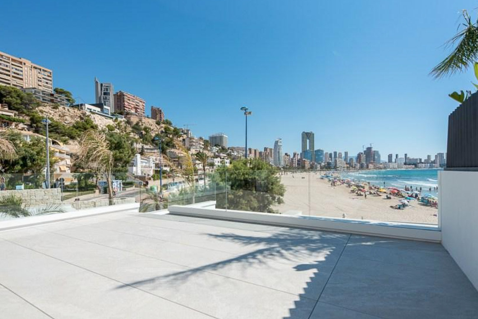 Terrasse extérieure avec carrelage grand format et garde-corps en verre, offrant une vue panoramique sur la plage et les gratte-ciel de Benidorm.