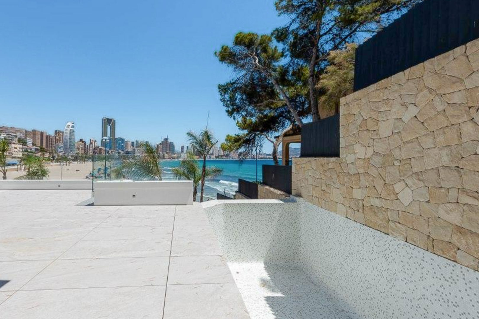 Terrasse extérieure avec dallage en pierre claire et piscine en mosaïque blanche, offrant une vue panoramique sur la mer et les gratte-ciels de Benidorm.