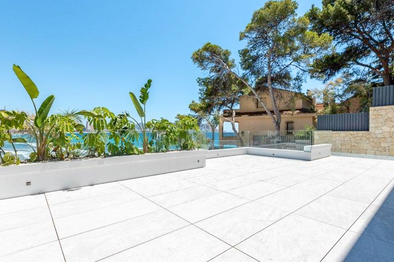 Terrasse extérieure avec revêtement en grès cérame grand format, garde-corps en verre sans cadre et jardinières intégrées offrant une vue sur la mer Méditerranée.