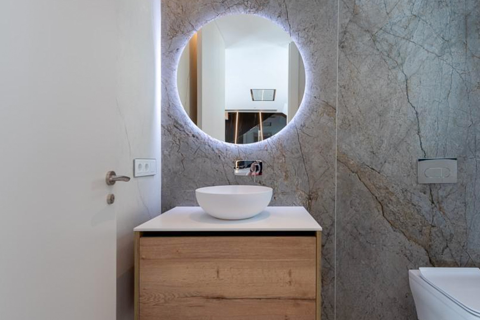 Toilettes d'invités avec carrelage mural effet marbre gris, miroir circulaire rétroéclairé et meuble vasque en bois avec robinetterie encastrée.