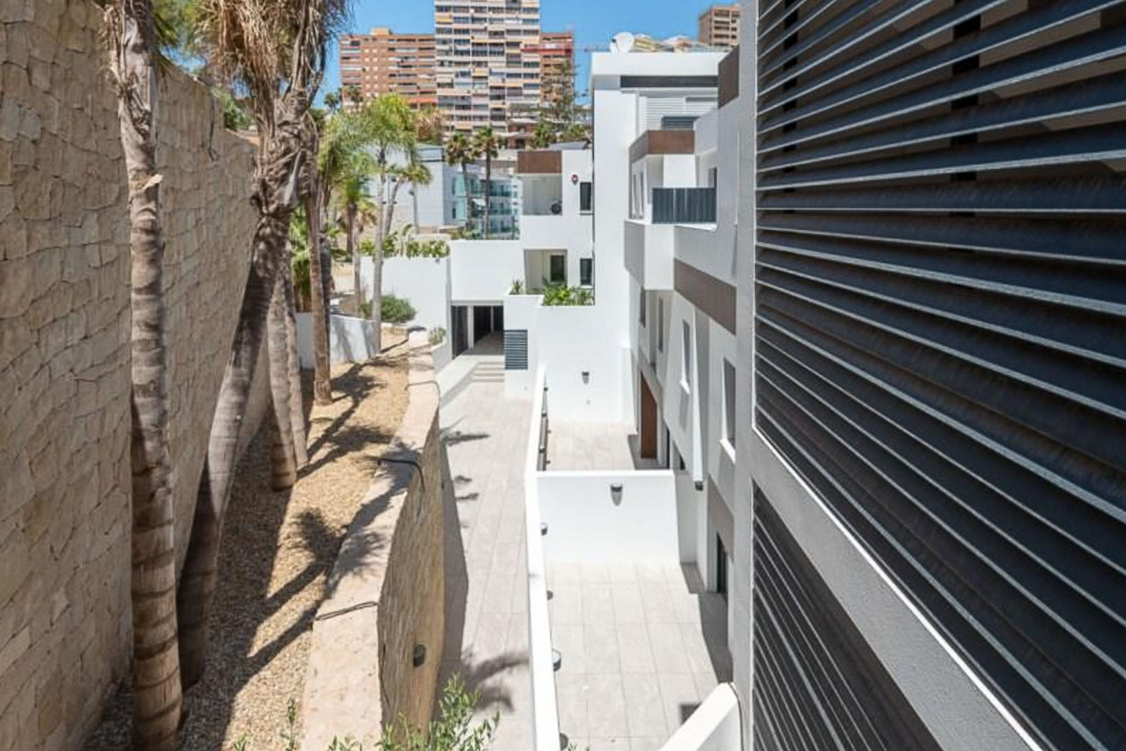 Vue extérieure du complexe avec façades blanches, mur en pierre naturelle et palmiers. L'architecture inclut des persiennes horizontales et des allées pavées.