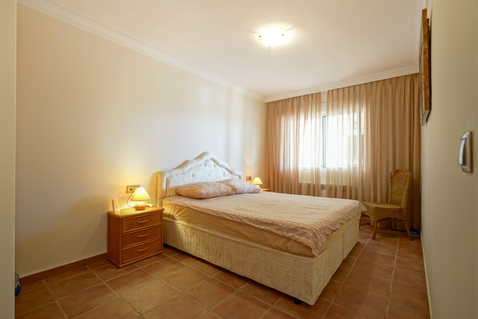Chambre à coucher avec sol en terre cuite, lit double à tête de lit capitonnée, table de chevet en bois et grande fenêtre avec rideaux voilages.