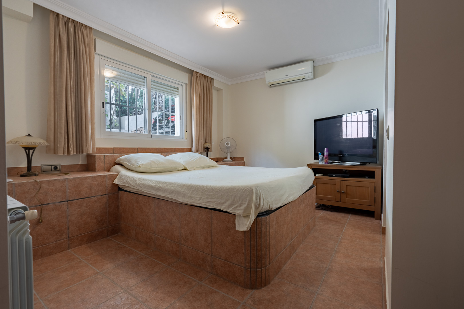 Chambre avec cadre de lit maçonné en grès cérame, climatisation réversible, radiateur et fenêtre offrant une luminosité naturelle constante.