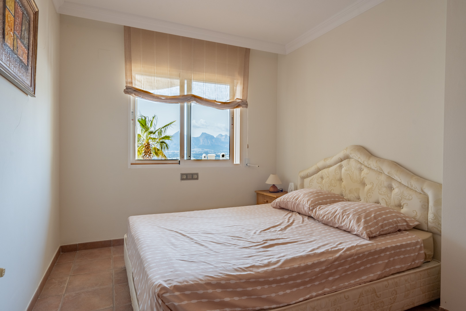 Chambre avec sol en terre cuite et tête de lit capitonnée. La fenêtre offre une vue sur les montagnes environnantes et un palmier extérieur.