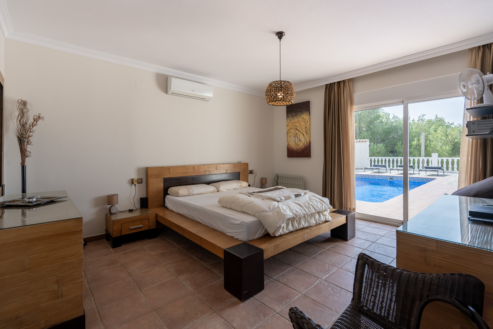 Chambre principale avec sol en terre cuite, climatisation et accès direct à la terrasse de la piscine par de grandes baies vitrées coulissantes.