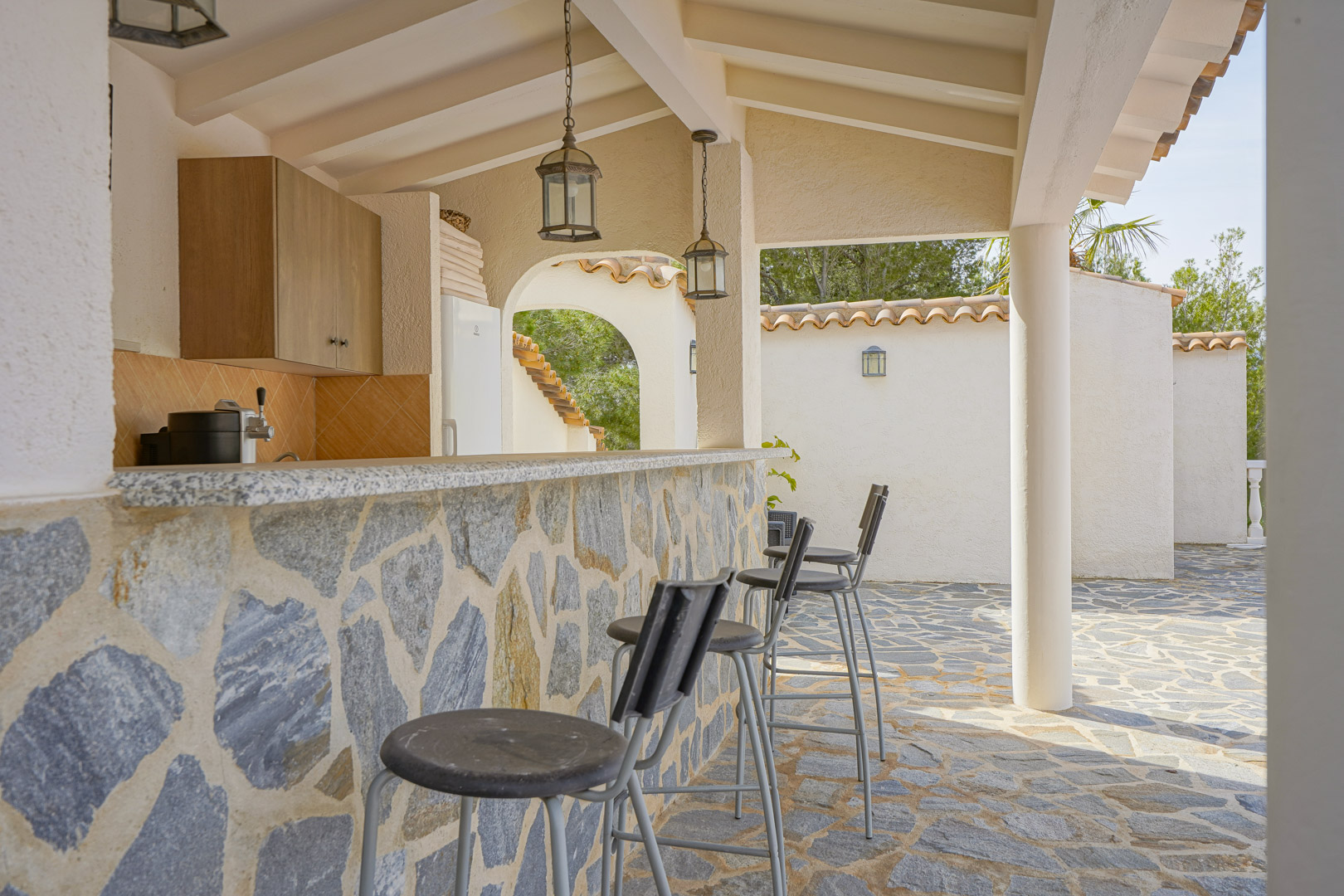 Cuisine d'été avec bar en pierre naturelle, plan de travail en granit et tabourets hauts, située sous un porche couvert aux poutres apparentes et lanternes suspendues.
