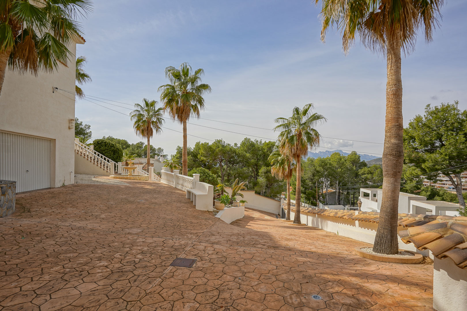 Entrée de la villa avec une large allée en béton imprimé, des palmiers matures et vue sur les montagnes. Comprend l'accès au garage et des balustrades blanches.