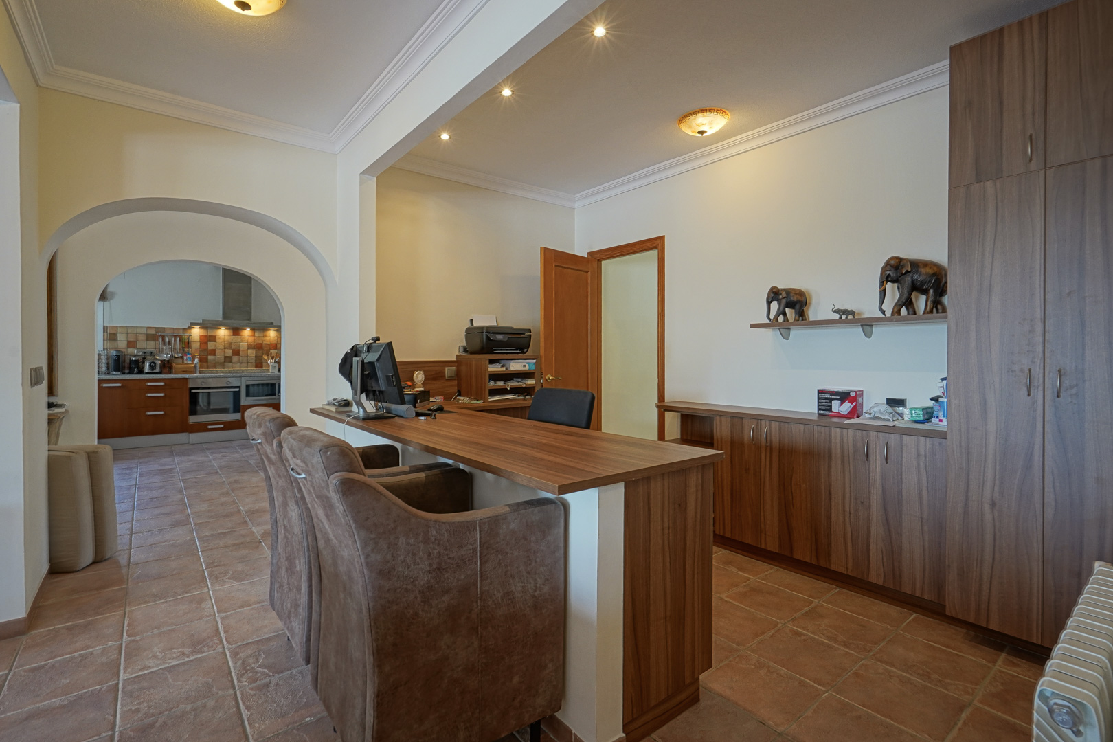 Espace bureau avec mobilier en bois sur mesure, sol en terre cuite et ouvertures voûtées offrant une vue sur la cuisine équipée.