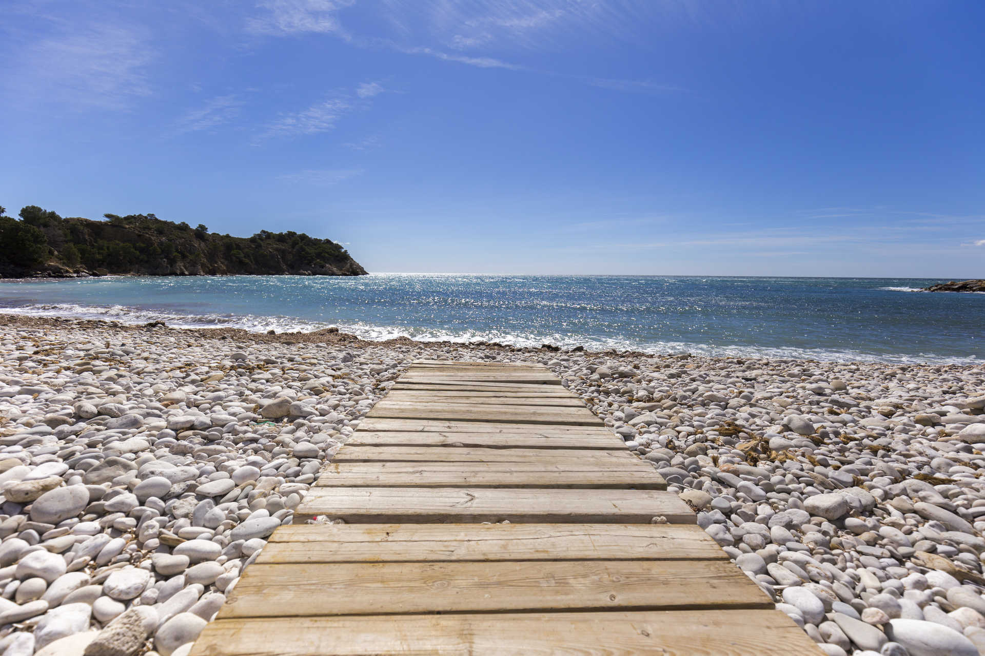 Plage de galets blancs à Altea avec une passerelle en bois menant aux eaux turquoise de la Méditerranée, entourée d'un paysage côtier naturel.