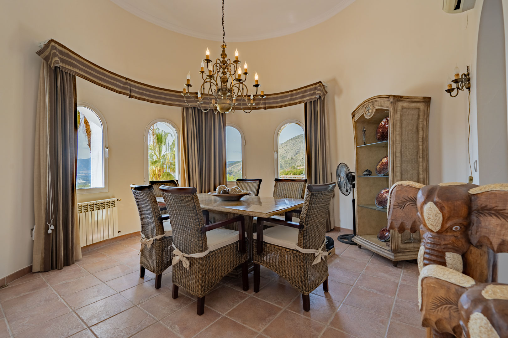 Salle à manger circulaire avec table en pierre, chaises en osier et lustre classique. Quatre fenêtres cintrées offrent une vue dégagée sur la Sierra de Bernia.