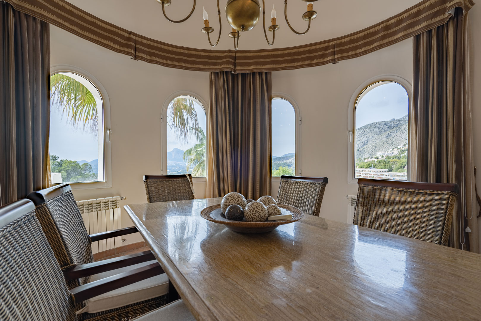 Salle à manger circulaire avec table en pierre polie et chaises en rotin. Quatre fenêtres cintrées offrent une vue panoramique sur la Sierra de Bernia et les montagnes d'Altea.