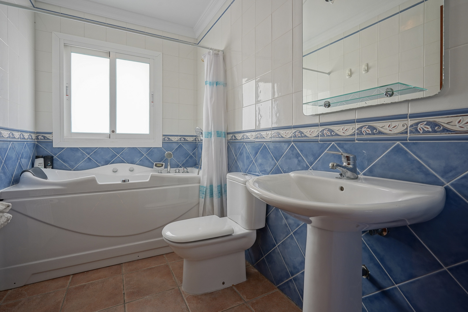 Salle de bain avec baignoire d'hydromassage, lavabo sur colonne et carrelage mural bleu et blanc. Sol en terre cuite et fenêtre apportant de la lumière naturelle.