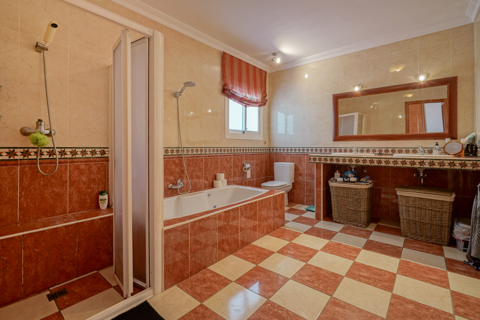 Salle de bain carrelée dans les tons crème et terre cuite, comprenant une baignoire encastrée, une douche séparée et un double lavabo sous un grand miroir.