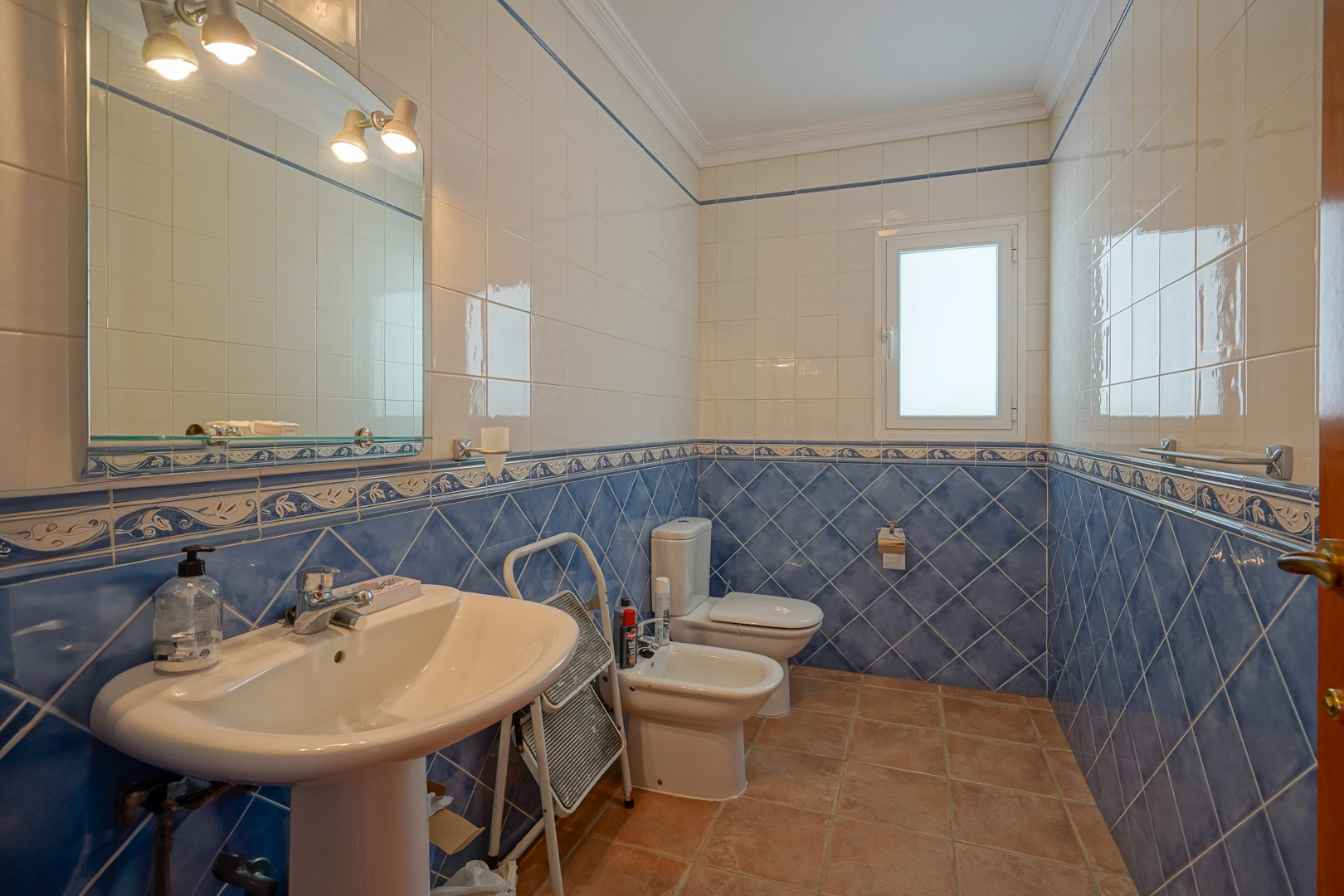 Salle de bains avec carrelage mural bleu et blanc, sol en terre cuite et sanitaires classiques comprenant un lavabo sur colonne, un bidet et un miroir avec éclairage.