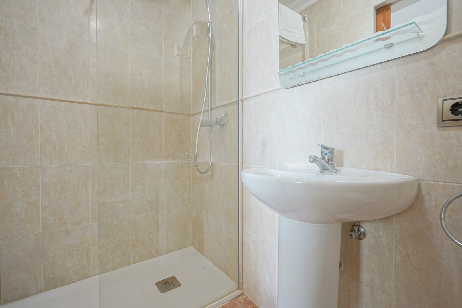 Salle de bains revêtue de carrelage beige, comprenant un lavabo sur colonne blanc et une douche à l'italienne avec paroi en verre et robinetterie chromée.