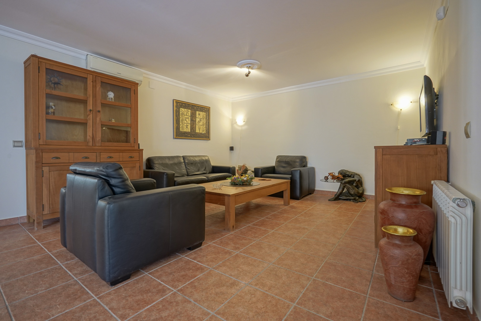 Salon avec canapés en cuir noir, mobilier en bois massif et sol en terre cuite. Équipé d'une climatisation réversible et de radiateurs de chauffage central.