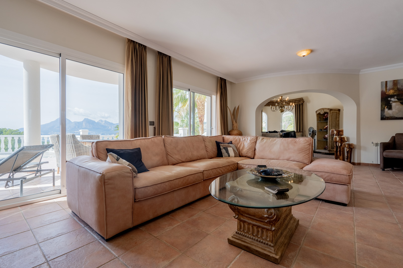 Salon lumineux avec sol en terre cuite, grand canapé d'angle en cuir et baies vitrées offrant une vue dégagée sur les montagnes et un accès à la terrasse.