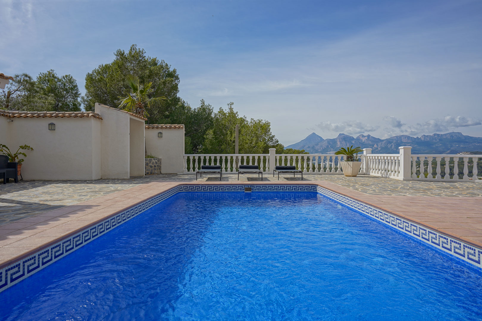 Terrasse de la piscine avec revêtement en pierre naturelle et balustrade blanche, offrant une vue panoramique sur les massifs du Puig Campana et de la Sierra Aitana.