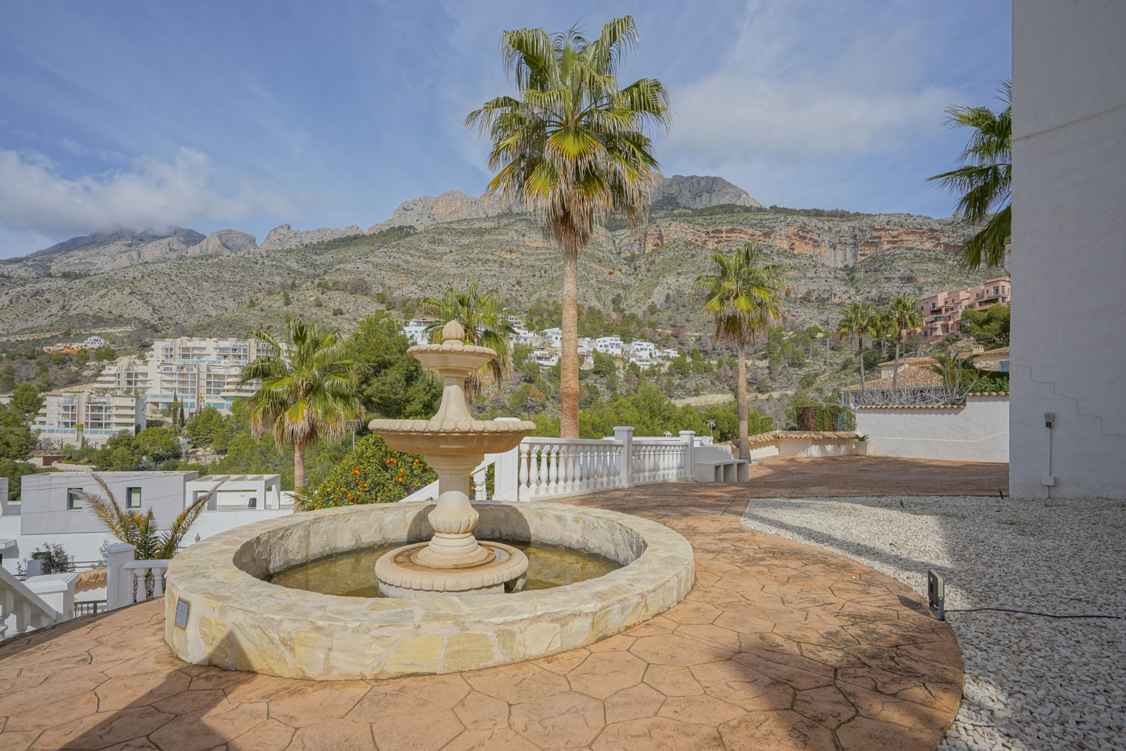 Terrasse extérieure avec fontaine en pierre sculptée et sol en béton imprimé, offrant une vue panoramique sur la Sierra de Bernia et ses palmiers.