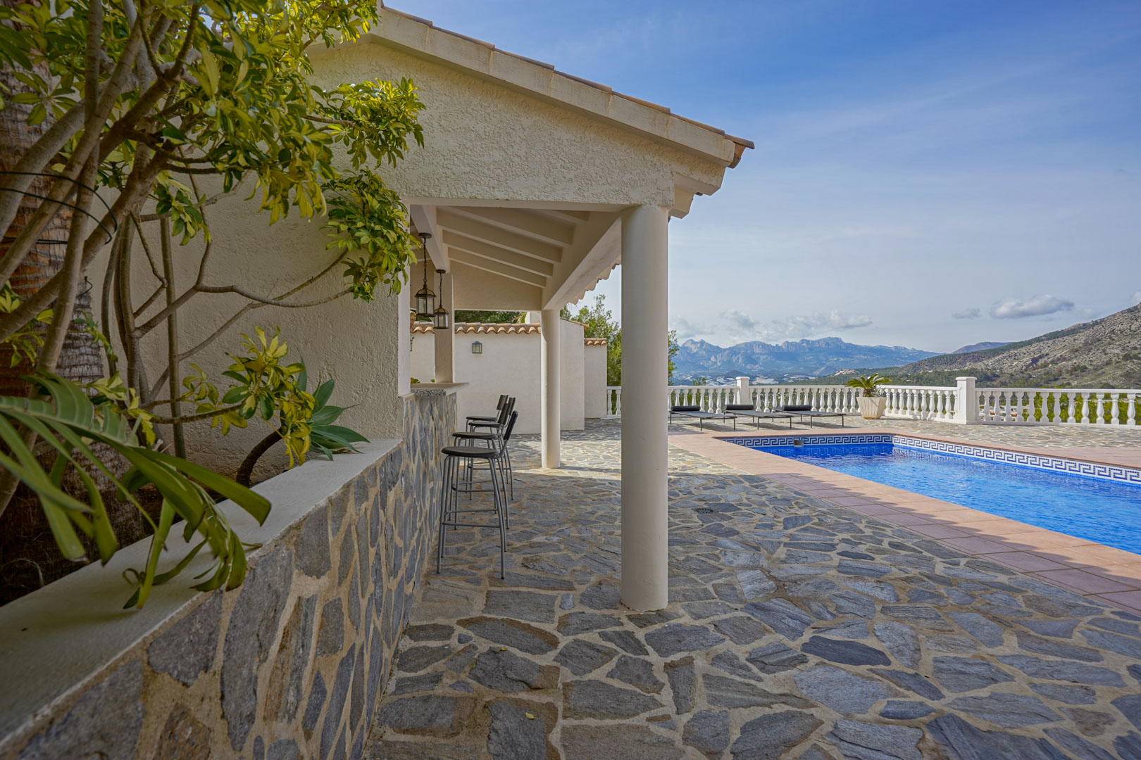 Terrasse extérieure en pierre naturelle avec cuisine d'été couverte, bar et piscine privée offrant une vue panoramique sur les montagnes d'Altea.