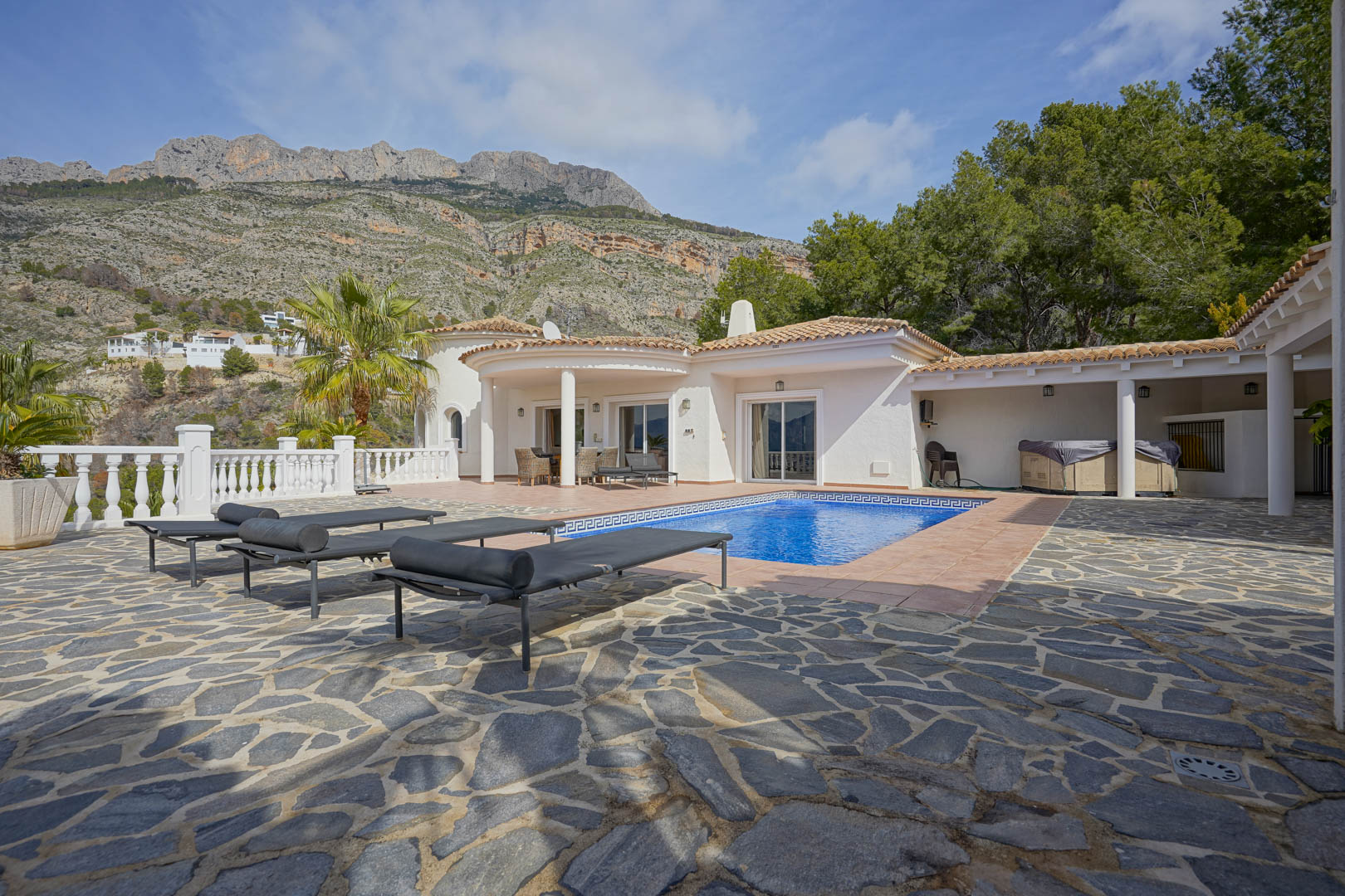 Terrasse principale avec piscine et dallage en pierre naturelle, offrant une vue dégagée sur la Sierra de Bernia et l'élégante façade blanche de la villa.