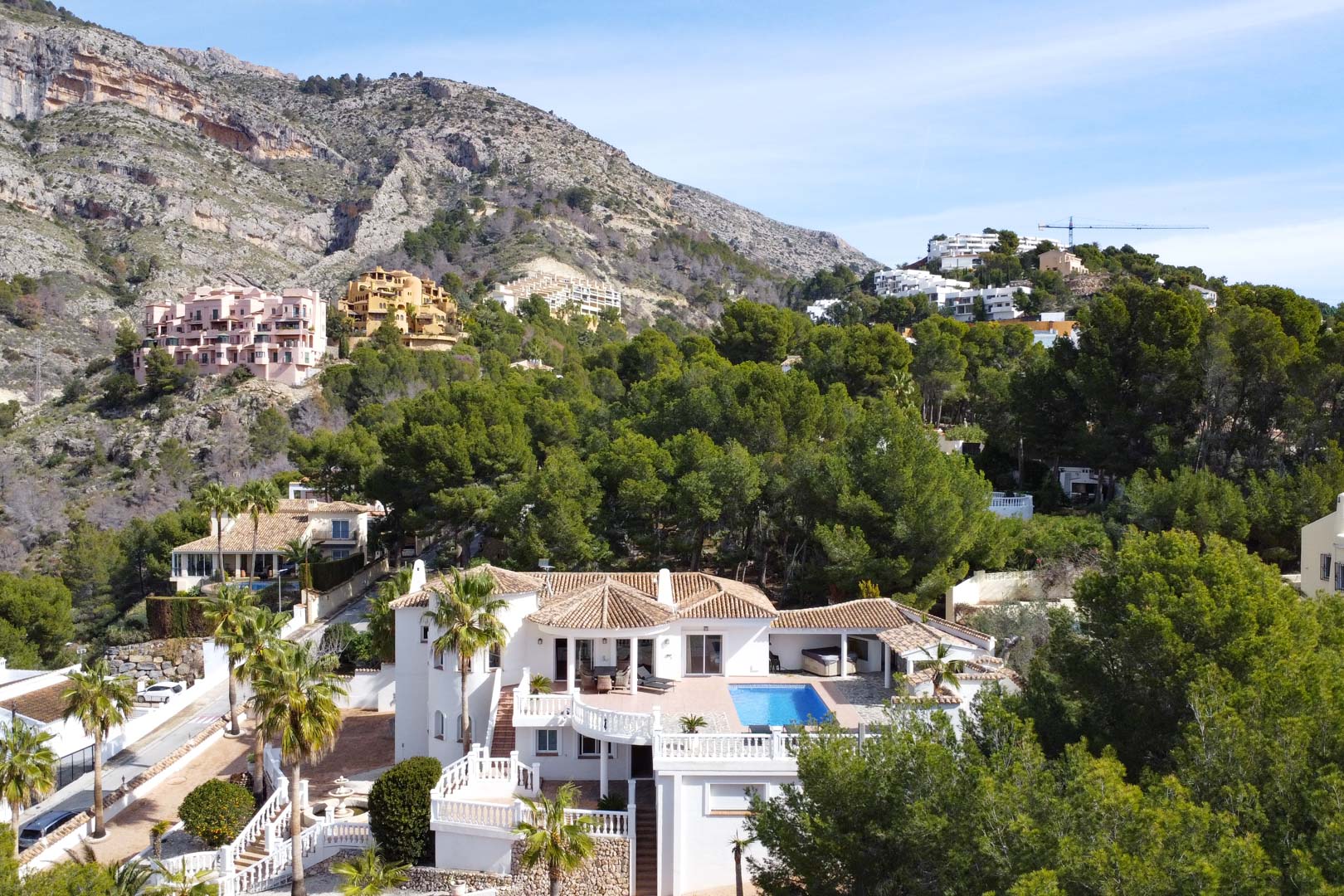 Vue aérienne de la villa avec toits en terre cuite, piscine privée et terrasses blanches, entourée de pins au pied de la Sierra de Bernia.