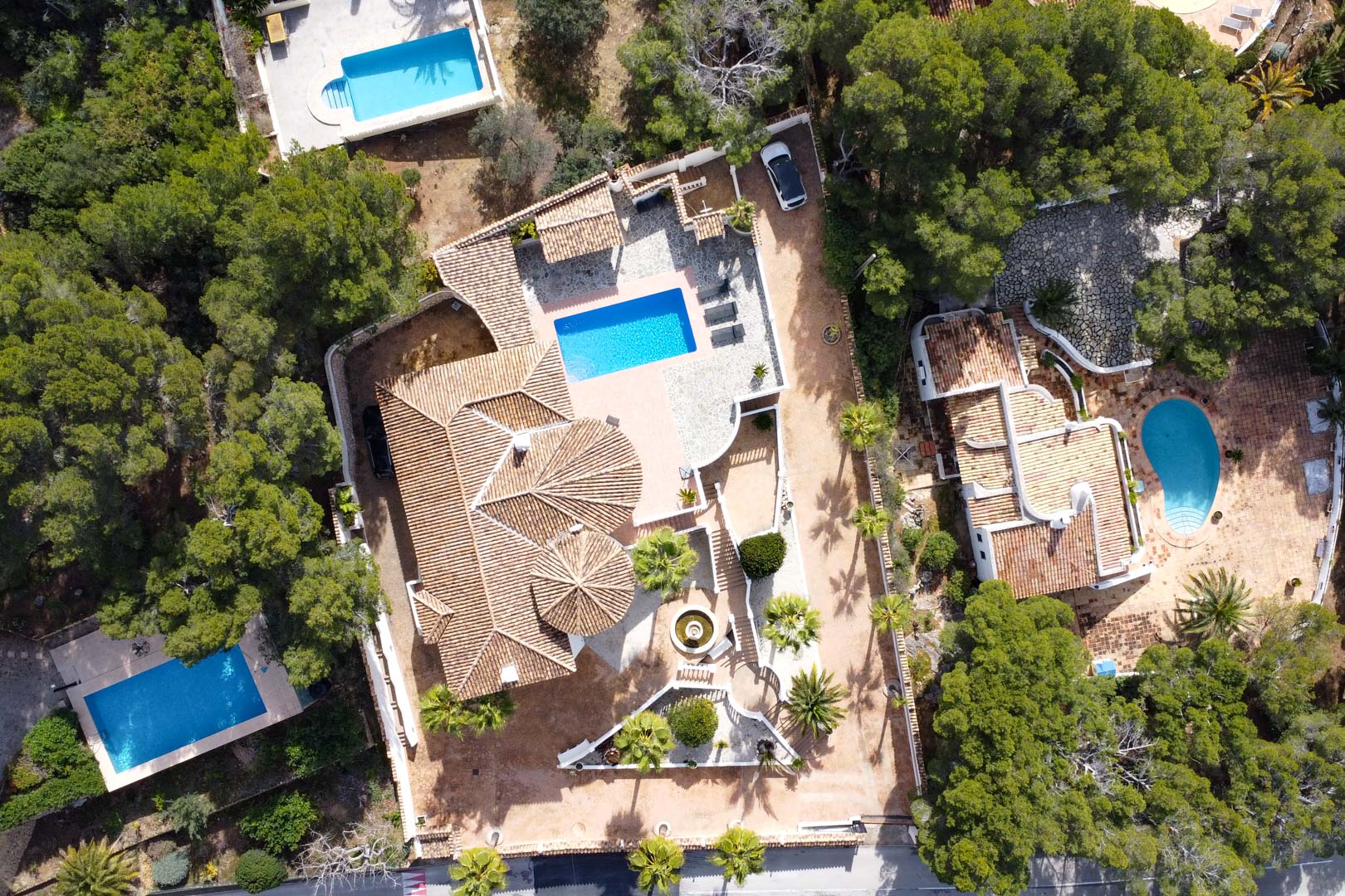 Vue aérienne de la villa soulignant l'architecture des toits en tuiles, la piscine rectangulaire et les terrasses en pierre entourées de pins et de palmiers.
