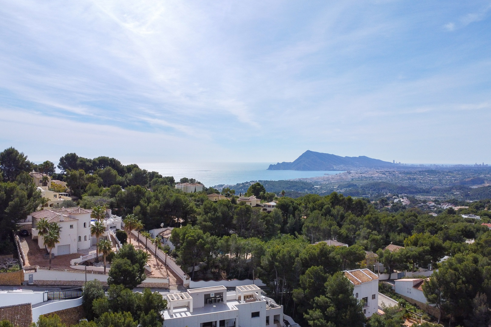Vue aérienne panoramique révélant la pinède environnante, les villas méditerranéennes et le littoral s'étendant jusqu'à la silhouette lointaine de Benidorm.