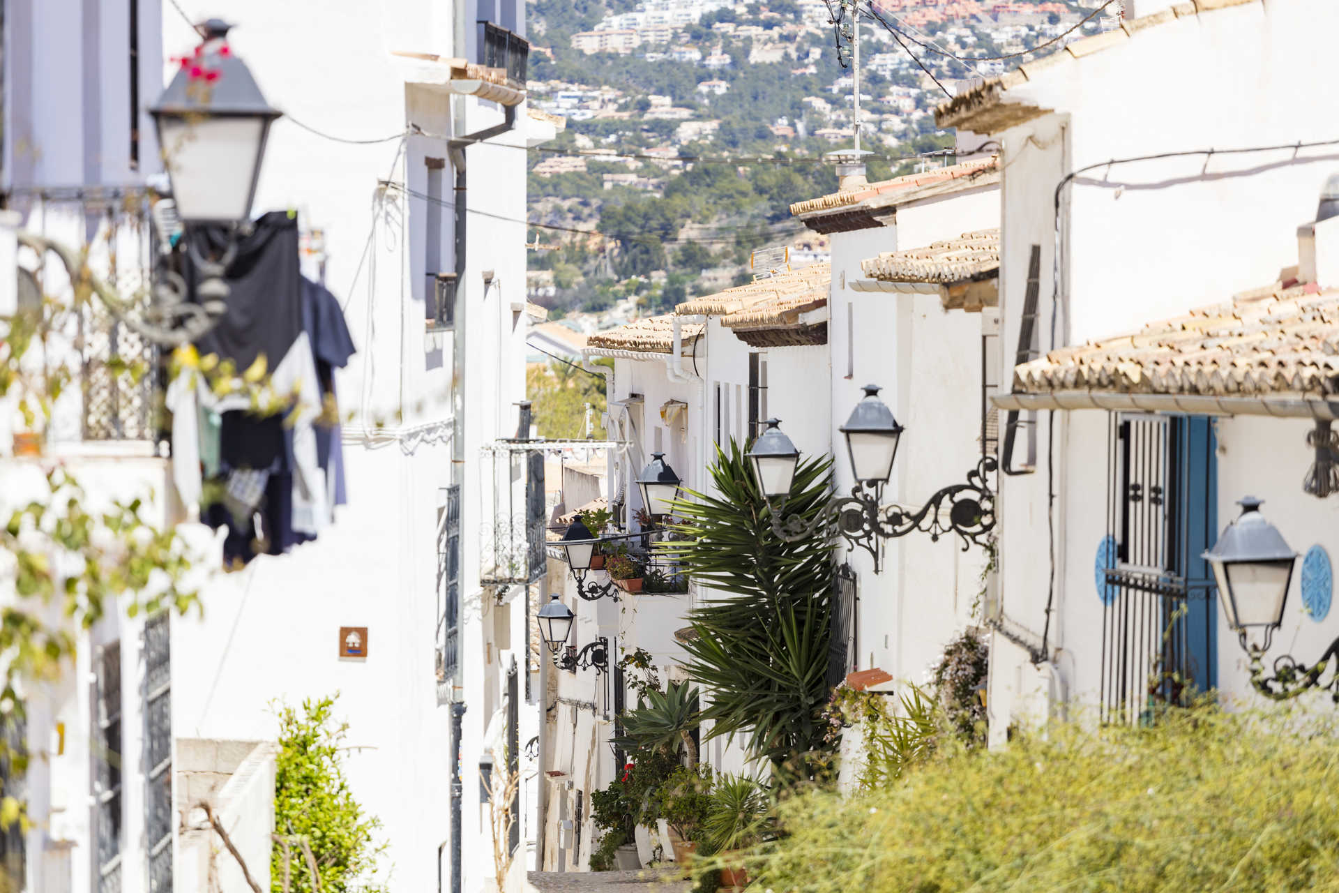 Vue d'une ruelle étroite dans la vieille ville d'Altea, avec des maisons blanches traditionnelles, des lanternes en fer forgé et une colline en arrière-plan.
