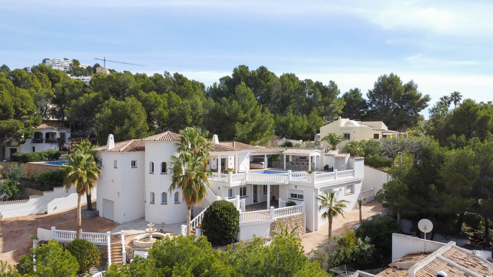 Vue extérieure de la villa méditerranéenne avec façade blanche, tour circulaire et terrasses à balustrades. Entourée de pins avec piscine au niveau supérieur.