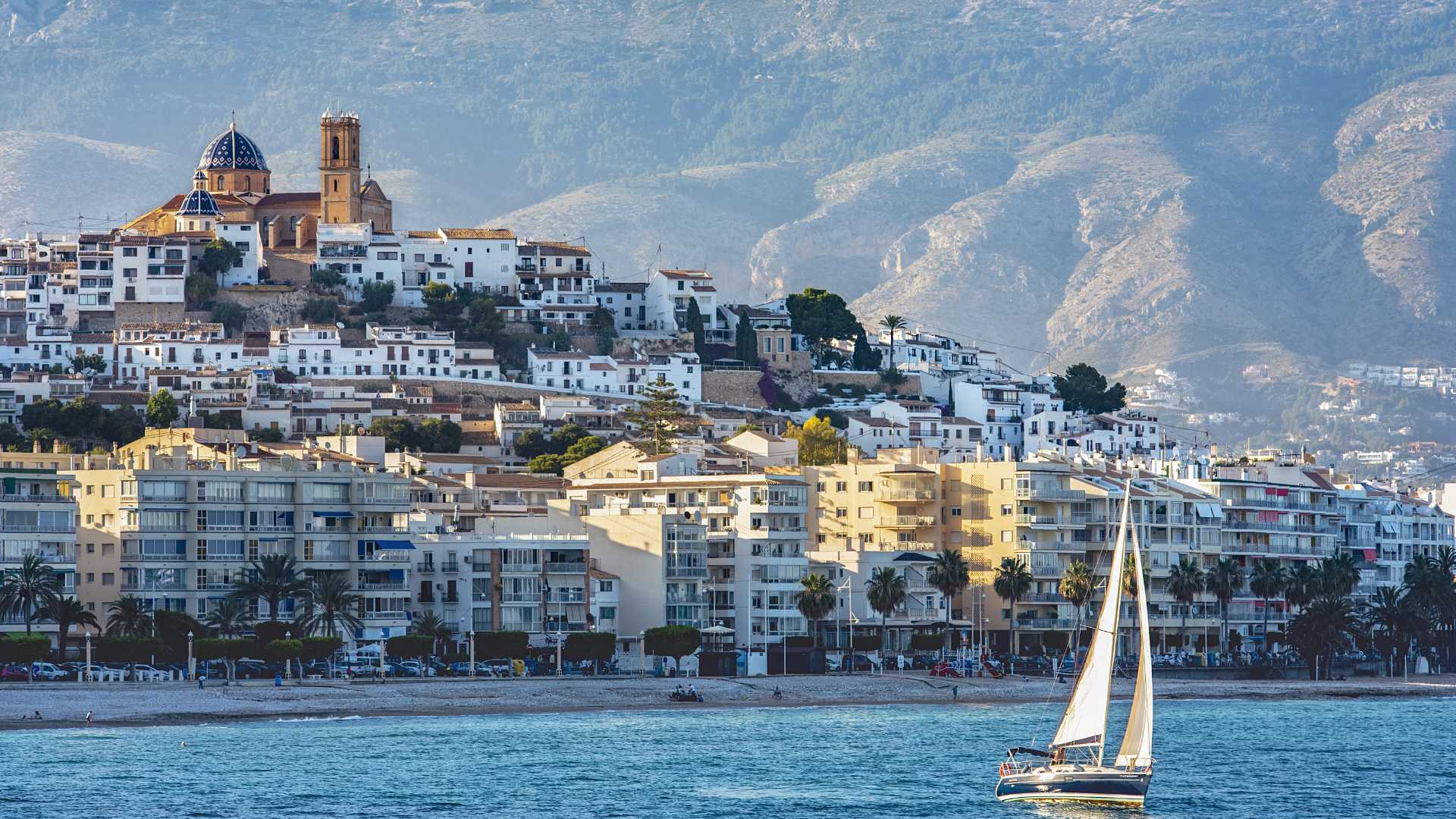 Vue panoramique de la vieille ville d'Altea avec son église aux coupoles bleues, surplombant la Méditerranée avec la Sierra de Bernia en arrière-plan.