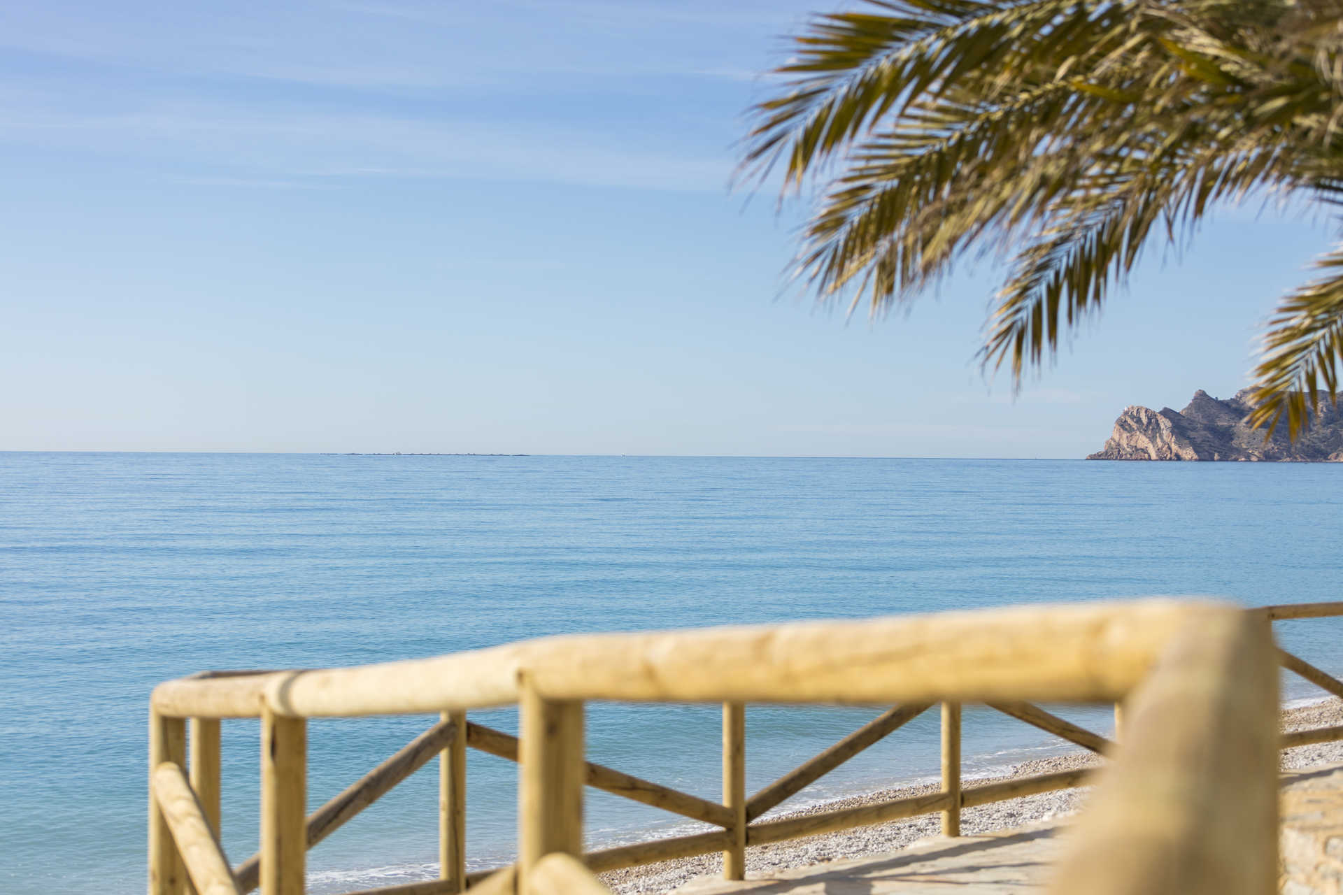 Vue panoramique sur la mer Méditerranée depuis le littoral d'Altea, avec des balustrades en bois, une plage de galets et des falaises rocheuses au loin.