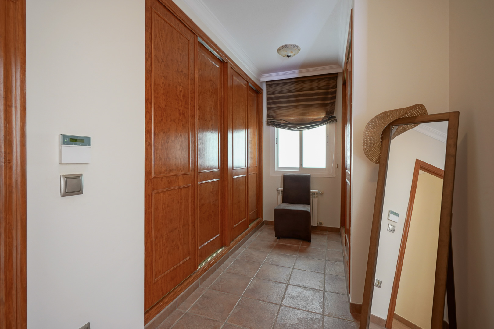 Zone de dressing avec de grands placards intégrés en bois, sol en terre cuite et fenêtre. Comprend un miroir sur pied et une chaise d'appoint.