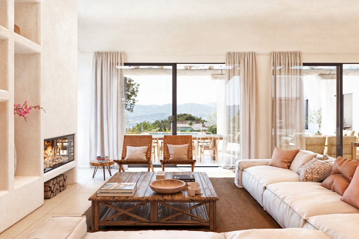 Salon lumineux avec cheminée intégrée, table basse en bois rustique et grandes baies vitrées offrant une vue panoramique sur la vallée et un accès à la terrasse.