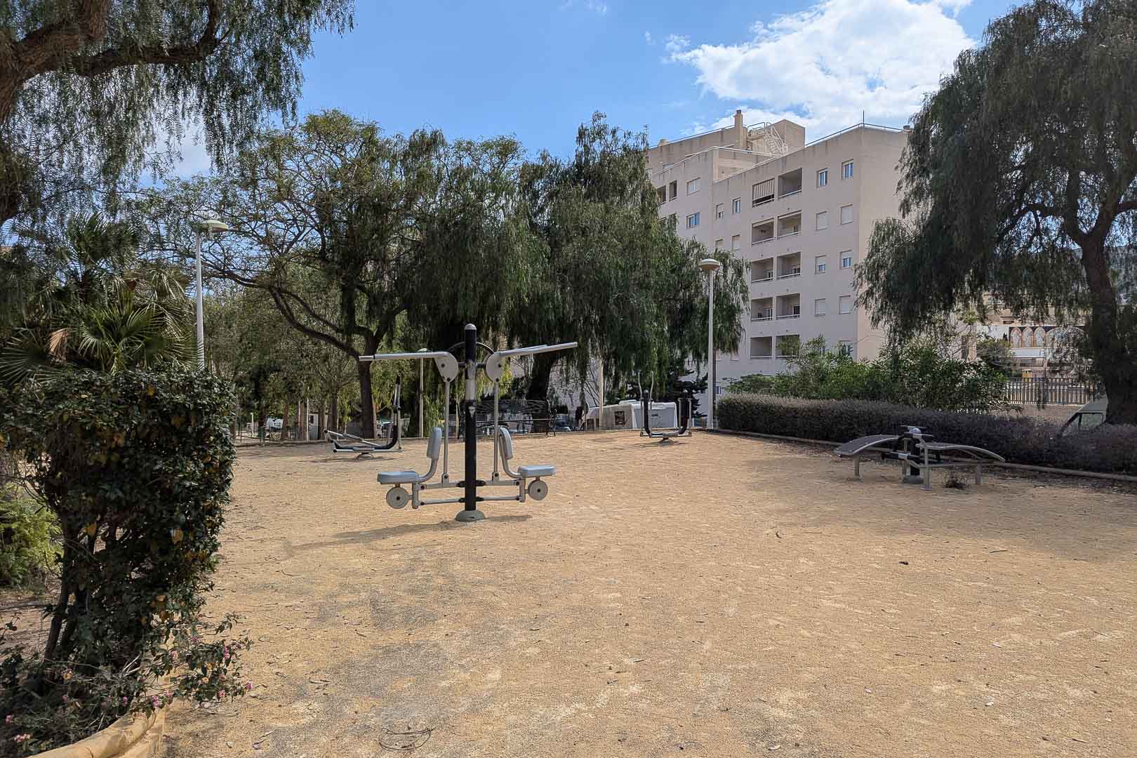 Espace de fitness en plein air équipé d'appareils de musculation, entouré d'arbres matures et de végétation dans un quartier résidentiel calme.