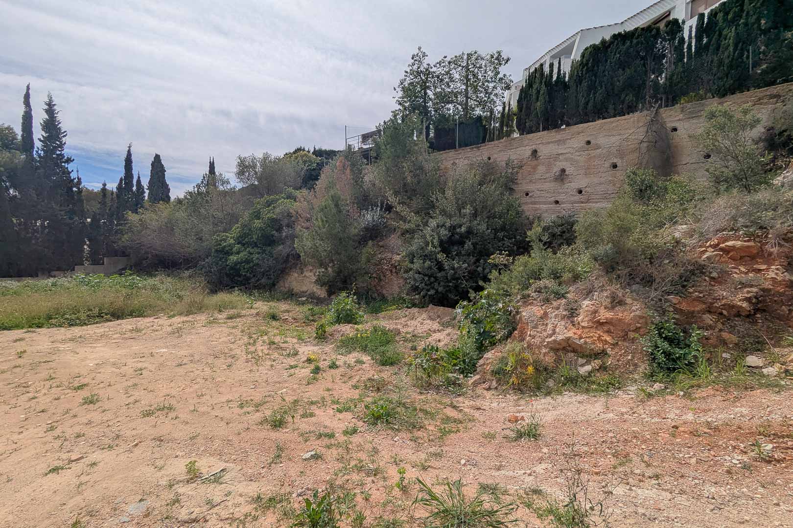 Terrain surélevé à La Corona avec mur de soutènement en béton et végétation méditerranéenne. Emplacement prêt pour le développement d'un projet de villa sur mesure.