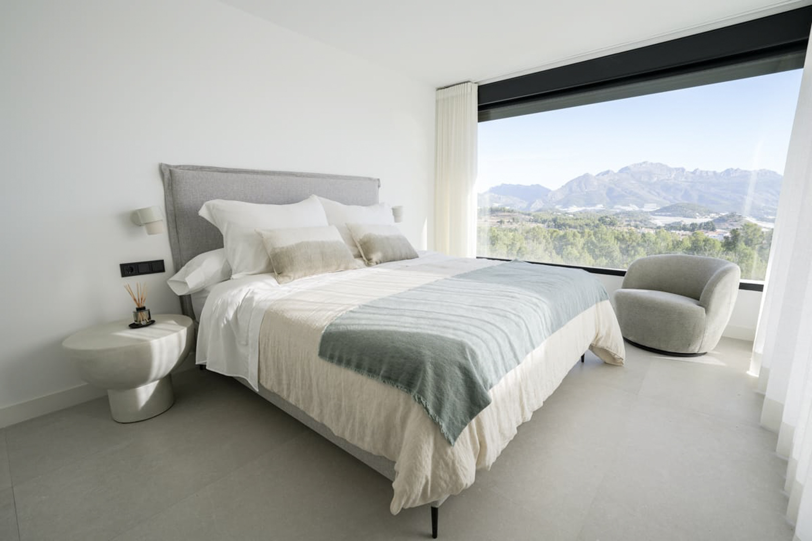 Chambre lumineuse avec lit double, fauteuil gris et vue sur la montagne à travers une grande fenêtre.