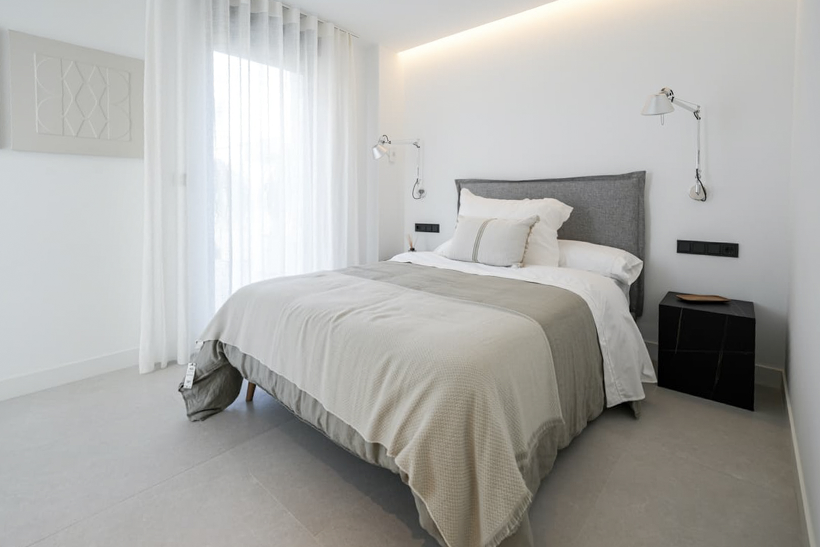Chambre lumineuse avec lit double, tête de lit rembourrée grise et lampes murales réglables. Sol en porcelaine.